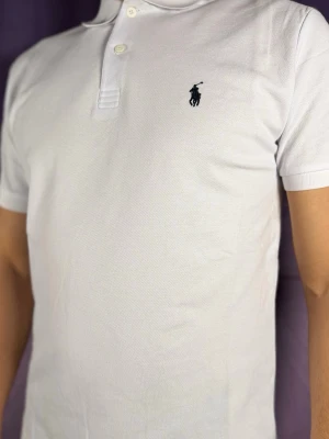 Vit pikétröja från Polo Ralph Lauren - Vit pikétröja från Polo Ralph Lauren med klassisk krage, korta ärmar och två knappar framtill. Ikonisk broderad logga på bröstet. Perfekt för en clean och stilren look.