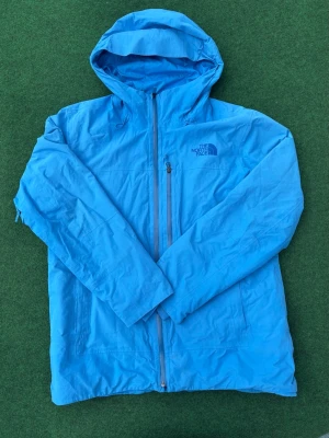 Blå vindjacka från The North Face - Snygg blå vindjacka från The North Face med huva och dragkedja framtill. Jackan har två sidofickor och logotyp på bröstet. Perfekt för blåsiga dagar. Storlek L   En metalldel på dragkedjan är av som bilden visar men den funkar som vanligt!! 