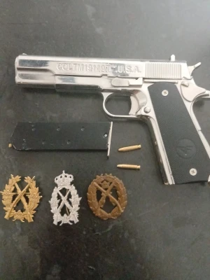 Metallisk Colt 1911A1 replika med märken - Säljer en metallisk replika av Colt 1911A1 med svart grepp, flera lösa patroner, ett magasin samt tre militära märken i guld, silver och brons. Perfekt för samlare eller dekoration. Pistolen har tydliga gravyrer och realistisk design. Pistolen går ej att skjuta med.