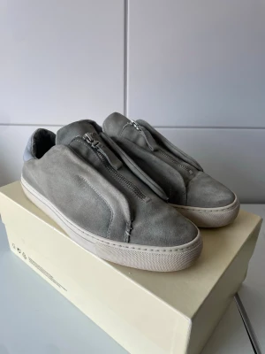 Gråa Axel Arigato skor med dragkedja  - Stilrena grå sneakers i mocka med dragkedja framtill istället för klassiska snören. Skorna har en vit sula och en mjuk, minimalistisk design. Perfekta för dig som gillar en clean look och vill ha något annorlunda än vanliga sneakers.