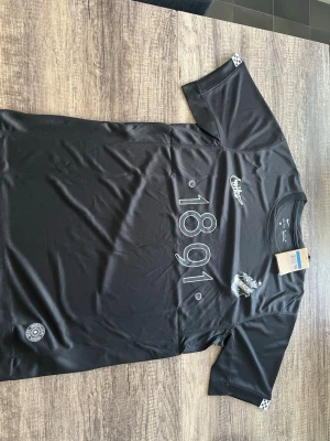Svart AIK smokinglirare  - Svart sportig t-shirt från Nike med AIK-logga och texten '1891' på bröstet. Tillverkad i Dri-Fit-material som andas, perfekt för träning eller match. Kortärmad modell med rund hals och diskreta detaljer på ärmen. Oanvänd!!! Fraktar dagen efter!!!