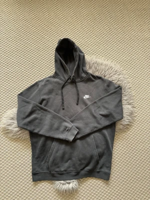 Svart hoodie från Nike - Svart hoodie från Nike med vit broderad logga på bröstet. Klassisk modell med huva och dragsnören samt känguruficka framtill. Mjuk bomullskänsla och ribbade muddar vid ärmslut och nederkant. Perfekt för en avslappnad streetstyle.