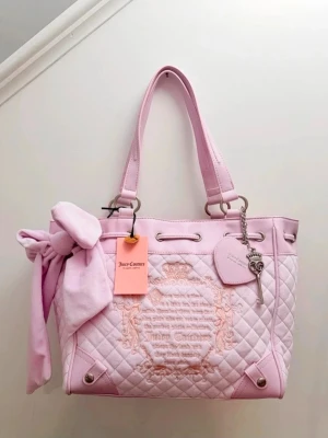  Juicy Couture daydreamer fairytale tote  - Superfin ljusrosa quiltad handväska från Juicy Couture med broderad logga framtill, dekorativ rosett på handtaget och silverfärgade detaljer. Väskan har två handtag, hjärtformad tag och nyckelcharm. Perfekt för dig som gillar en söt och trendig look.
