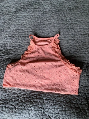 Rosa/orange spetsdetaljerad linnetopp - Söt rosa/orange linnetopp med broderade detaljer och spets längs axelpartiet. Toppen är ärmlös och har en hög halsringning med smala axelband. Perfekt för varma dagar och ger en romantisk vibe till din outfit. En söm har gått upp efter en tvätt.