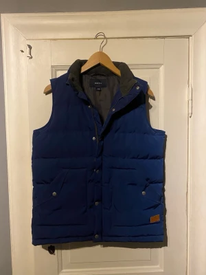 Blå dunväst från GANT - Snygg blå dunväst från GANT med hög krage och tryckknappar framtill. Västen har två stora fickor med lock och en liten läderdetalj på ena fickan. Perfekt för lager-på-lager och chill stil. Fodrad insida i mörkare nyans.