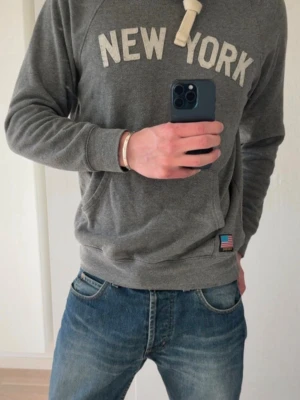 Witty Knitters Hoodie med New York-tryck - Säljer en grå hoodie med texten 'NEW YORK' i vitt på bröstet och en liten amerikansk flagga vid nederkanten från Witty Knitters. Hoodien har huva med breda vita snören och ribbade muddar vid ärmar och midja. Perfekt för dig som gillar streetwear och enkel stil.