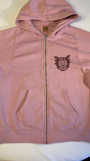 Rosa hoodie från Uzi Made - Säljer en rosa hoodie från Uzi Made med dragkedja framtill och ett brunt tryck med logga på bröstet. Hoodien har huva och kängurufickor. Tillverkad i mjukt bomullsmaterial, perfekt för en chill och avslappnad stil.