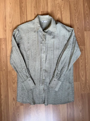 Grön skjorta med knyt bak från Cubus - Stilren grön skjorta från Cubus med lång ärm och knappar framtill. Skjortan har en unik struktur i tyget och två knytband på ryggen för extra detalj. Perfekt för dig som gillar en avslappnad men ändå trendig look.