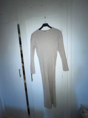 Beige ribbad långklänning - Långärmad beige långklänning i ribbad stickad design. Klänningen har rund halsringning och en kroppsnära passform som framhäver siluetten. Perfekt för dig som gillar en stilren och enkel look.