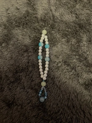 Handgjord pärlberlock i blått och vitt - Unik handgjord berlock med vita och blå pärlor i olika storlekar. Pärlorna är trädda på en genomskinlig tråd och bildar en droppformad dekoration. Perfekt att hänga på väska, mobil eller nyckelring för en personlig touch.