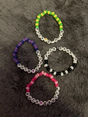 Pärlarmband Fuck Cancer 4 pack - Färgglada pärlarmband med texten 'FUCK CANCER' i vita bokstavspärlor. Finns i olika färgkombinationer: rosa, lila, svart/vit och grön/gul. Elastisk tråd och plastpärlor. Perfekt statement-accessoar för att visa stöd och attityd.