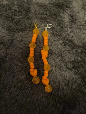Nyckelring med orange pärlor och hjärtan 2 pack - Handgjort nyckelring med orange plastpärlor och genomskinliga facetterade pärlor. Nyckelring har även små hjärtformade pärlor som ger en lekfull touch. Perfekt för dig som gillar färgglada accessoarer och vill sticka ut. Ena med guld detalj, andra med silver. 