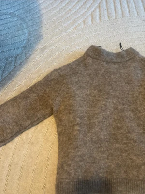 Beige stickad tröja med hög krage - Mysig beige stickad tröja med lite höjd krage och långa ärmar. Tröjan har en enkel och stilren design med ribbstickade muddar vid ärmslut och nederkant. Perfekt för kyliga dagar och lätt att matcha med olika outfits.