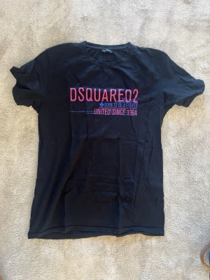 Svart DSQUARED2 t-shirt med tryck - Svart t-shirt från DSQUARED2 med rosa och lila tryck framtill där det står 'DSQUARED2' och 'UNITED SINCE 1964'. Klassisk rund hals och korta ärmar. Perfekt för dig som gillar streetwear och vill ha något stilrent men ändå med attityd.