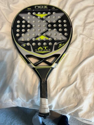 NOX AT Genius Junior padelracket - Säljer ett NOX AT Genius Junior padelracket i svart, grått och gult. Racketen har rund form med många hål och ett stort X-mönster på träffytan. Handtaget är lindat med vitt greppband. Perfekt för dig som vill ta ditt padelspel till nästa nivå.