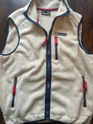 fleeceväst från Patagonia - Snygg beige fleeceväst från Patagonia med marinblå detaljer och dragkedjor. Västen har tre fickor med dragkedja och röda zip-pullers. Perfekt för lager-på-lager och chill stil. Mjuk och varm med hög krage och klassisk Patagonia-logga på bröstet.
