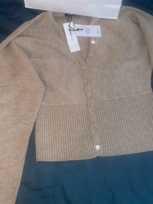 Beige ribbad kofta från Gina Tricot - Snygg beige kofta från Gina Tricot med ribbad nederkant och knappar framtill. Koftan har v-ringning och långa ärmar. Materialet består till 51% av GRS-certifierad återvunnen polyester, vilket gör den både trendig och hållbar.
