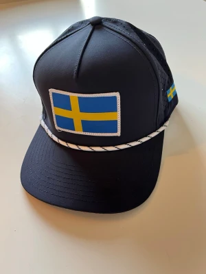 Sverige keps med band (Golfio) - Svart keps med böjd skärm och ventilerande meshpaneler på sidorna. Stor svensk flagga framtill och en mindre flagga på sidan. Vit och svart snodd längs skärmens kant. Perfekt för dig som vill visa din svenska stil.