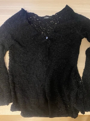 Svart topp CHIMI - Svart topp från CHIMI med grovstickad struktur och v-ringning. Koftan har en knapp framtill och långa ärmar. Perfekt för lager-på-lager och ger en cool, avslappnad vibe till din outfit.
