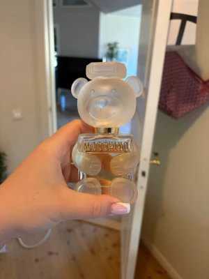 Moschino Toy 2 parfym björnflaska - Unik parfym från Moschino i en genomskinlig glasflaska formad som en björn. Flaskan har frostad finish på detaljerna och guldiga inslag vid halsen. Perfekt för dig som gillar lekfull och trendig design på parfymflaskor.