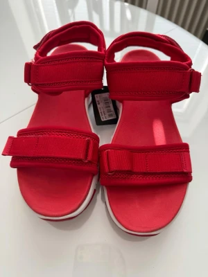 Nya Röda sportiga sandaler med kardborre - Snygga röda sandaler med vita sulor och grovt mönster undertill. Justerbara kardborreremmar över foten och runt hälen för perfekt passform. Perfekta för en aktiv stil och riktigt bekväma att gå i. Nypris 699:- 