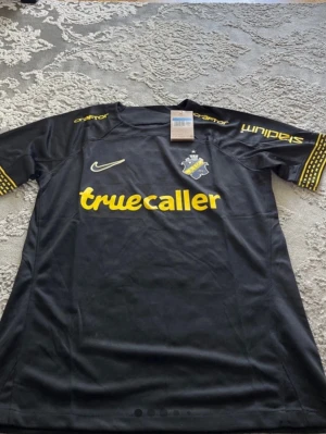 Svart AIK matchtröja Nike - Säljer en svart AIK matchtröja från Nike med klubbmärke på bröstet och gula detaljer. Tryck med 'truecaller' på magen och sponsorloggor på ärmarna. Tillverkad i lätt och ventilerande funktionsmaterial, perfekt för fotboll eller träning.