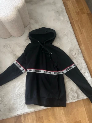 Svart hoodie från Moschino - Svart hoodie från Moschino med vit och röd logoband-detalj över bröstet och armarna. Klassisk huva med dragsko och ribbade muddar. Perfekt för en trendig och avslappnad streetstyle-look.