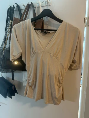 Beige v-ringad topp  - Trendig beige topp med v-ringning och korta puffärmar. Toppen har rynkade detaljer vid sidorna och en figurnära passform🥰