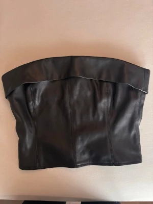 Svart axelbandslös skinnkorsett - Snygg svart axelbandslös korsettopp i skinnimitation med bred kant upptill och dragkedja bak. Perfekt för att skapa en edgy look och passar till både jeans och kjol. Stilren design utan extra detaljer för en clean vibe.