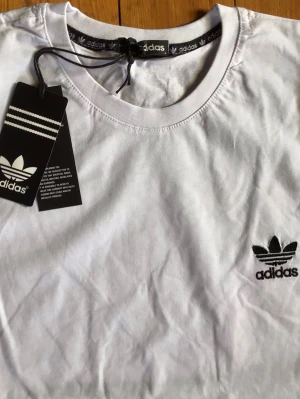 Vit Adidas t-shirt med logga - Vit t-shirt från Adidas med klassisk svart logga på bröstet. T-shirten har rund halsringning och korta ärmar. Tillverkad i mjuk bomull för en skön känsla. Perfekt för en sportig och avslappnad stil.
