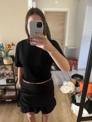 Svart volangkjol i satin - Snygg svart kortkjol med volangkant och lätt glansig satinfinish. Kjolen har en figurnära passform och är perfekt för dig som gillar en trendig och stilren look. Enkel att matcha med olika toppar och accessoarer.