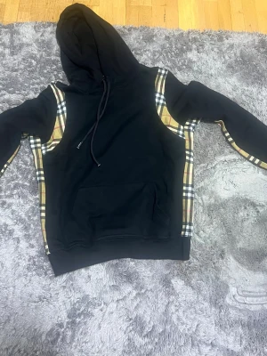 Svart hoodie med rutiga detaljer - Svart hoodie med klassiska Burberry-rutiga paneler längs sidorna och ärmarna. Stor huva med dragsko och en stor magficka framtill. Hoodien har en avslappnad passform och är tillverkad i mjukt material, perfekt för en trendig streetwear-look.