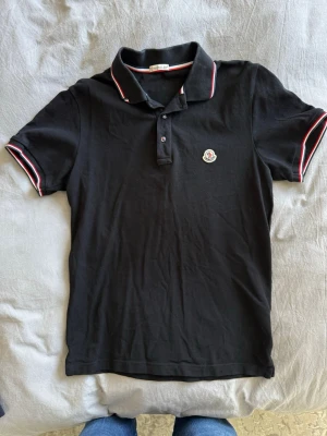 Moncler pikétröja svart - Svart pikétröja från Moncler med klassisk krage och knäppning framtill. Tröjan har röda och vita ränder på krage och ärmslut samt Moncler-logga på bröstet. Perfekt för en stilren och sportig look.