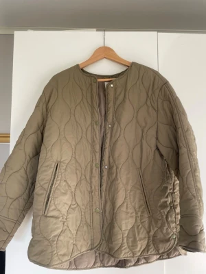 Beige quiltad jacka med tryckknappar - Quiltad jacka i beige med rund halsringning och tryckknappar framtill. Jackan har två fickor och ett vågigt mönster i sömmarna. Oversized passform och lätt vadderad, perfekt för lager-på-lager. Materialet ser ut att vara polyester eller bomullsblandning.