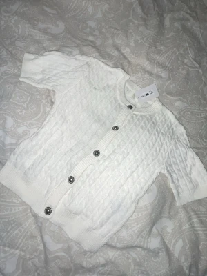 Vit kabelstickad kofta från H&M - Snygg vit kabelstickad kofta från H&M med korta ärmar och ribbade muddar. Koftan har rund halsringning och silverfärgade knappar framtill. Perfekt att slänga över en topp för en clean och fräsch look.