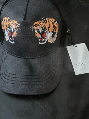 Svart GG keps med tigertryck - Svart keps  med två coola tigerhuvuden framtill och diskret GG-mönster i tyget. Justerbar rem med klassiska gröna och röda ränder. Tillverkad i bomull och polyester. Perfekt statement-piece för dig som vill sticka ut.