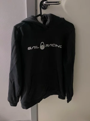 Svart hoodie från Sail Racing - Svart hoodie från Sail Racing med vit logga tryckt på bröstet. Klassisk modell med huva och stor magficka framtill. Perfekt för en avslappnad och sportig stil. Tillverkad i mjukt material för extra komfort.