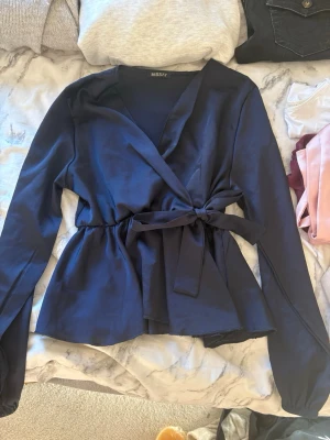 Mörkblå omlottblus från Missäy - Snygg mörkblå omlottblus från Missäy med knytband i midjan och långa ärmar. Blusen har en feminin siluett och är tillverkad i ett mjukt, lätt material som ger ett elegant fall. Perfekt för att lyfta vilken outfit som helst.
