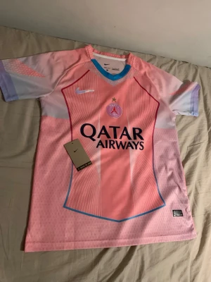 PSG rosa matchtröja Nike Dri-FIT - Säljer en rosa PSG matchtröja från Nike med Dri-FIT-material. Tröjan har ljusblå detaljer vid kragen och ärmarna, samt Qatar Airways-logga på bröstet. Perfekt för fotbollsträning eller att bära som supporter. PSG-emblem och Nike-logga framtill.