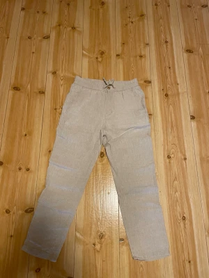 Beige Skagen Clothing linnebyxor med dragsko - Snygga beige linnebyxor med rak passform och elastisk midja med dragsko. Byxorna har fickor fram och bak, och är tillverkade i ett luftigt linnematerial som passar perfekt för varma dagar. Stilrena och enkla att matcha med olika toppar.