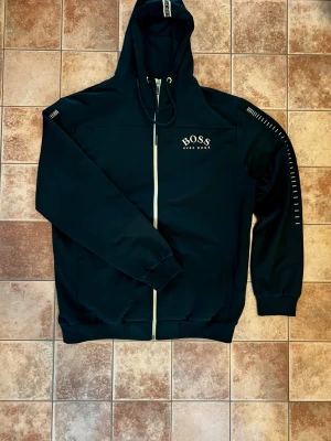 Svart hoodie från Hugo Boss - Svart hoodie från Hugo Boss med gulddetaljer och dragkedja framtill. Märkets logga i gult på bröstet och dekorativa linjer på ena ärmen. Huva med dragsko och två fickor framtill. Perfekt för en stilren och avslappnad look.