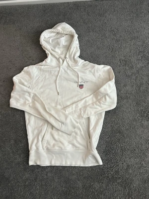 Vit hoodie från GANT med logga -  Vid hoodie från GANT med broderad logga och sköld på bröstet (Vänstra sidan). Hoodien har dragsko i huvan och en stor magficka framtill. Tillverkad i mjukt polyester material. Perfekt för en avslappnad och stilren look, som pass bra till Stocholm stilen. Storlek M