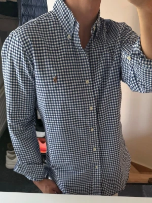 Blårutig skjorta från Ralph Lauren - Klassisk blårutig skjorta från Ralph Lauren med button-down krage och broderad logga på bröstet. Skjortan har långa ärmar, vita knappar och är tillverkad i mjuk bomull. Perfekt för en stilren och avslappnad look.lite missfärgad annars bra skick 
