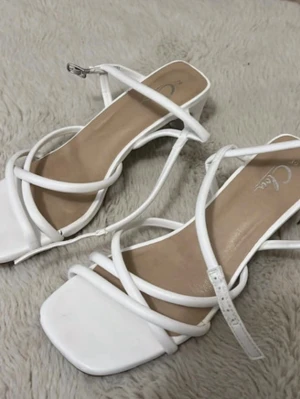 Vita strappy sandaler med klack - Säljer ett par vita sandaler med fyrkantig tå och tunna remmar i kors. Sandalerna har en medelhög klack och justerbart spänne vid ankeln. Snygg minimalistisk design som passar till många olika outfits.