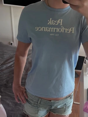 Ljusblå Peak Performance t-shirt - Ljusblå t-shirt från Peak Performance med vit text.  Klassisk rund halsringning och korta ärmar. Perfekt för en avslappnad och stilren look.  Den är i storlek 160 men passar oxå 170