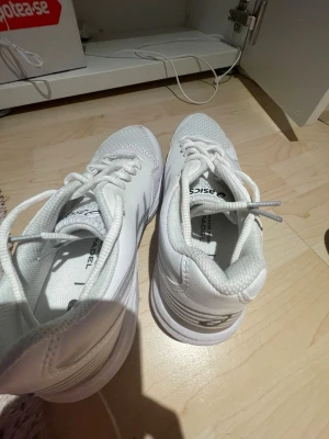 Vita Asics sneakers med silverdetaljer - Snygga vita sneakers från Asics med klassiska silverfärgade detaljer på sidorna. Skorna har en låg profil, snörning och ovandel i mesh och syntet för bra andningsförmåga. Perfekta för dig som vill ha en clean och sportig look.