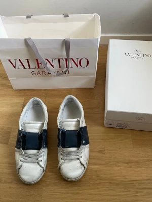 Valentino Garavani vita sneakers - Snygga vita sneakers från Valentino Garavani med bred svart rem över vristen. Skorna har klassisk låg modell, rund tå och vita snören. Tillverkade i läder med märkets logga på plösen. Perfekta för dig som gillar exklusiv streetstyle.