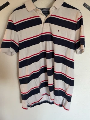 Randig pikétröja från Tommy Hilfiger - Klassisk pikétröja från Tommy Hilfiger med breda marinblå, röda och vita ränder. Tröjan har korta ärmar, krage och knäppning framtill. Liten logga broderad på bröstet. Perfekt för en avslappnad och stilren look.