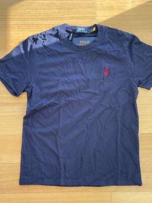 Marinblå Ralph Lauren T-shirt - 👕 Märke: Polo Ralph Lauren 📏 Storlek: S 🎨 Färg: Marinblå med röd logga ✨ ☀️ Fräsch och stilren t-shirt från Polo Ralph Lauren – perfekt för sommaren och lätt att matcha med både shorts och jeans. Tillverkad i mjuk bomull med klassisk passform och ikonisk röd logga på bröstet.  ✨ Skick: Helt ny med tags kvar 🧼 Aldrig använd 👌 Enkel, tidlös och riktigt snygg till vardags