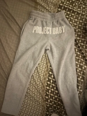 Grå mjukisbyxor Project Baby - Säljer ett par grå mjukisbyxor från Project Baby med bred resår i midjan och vita broderade bokstäver framtill. Byxorna har avslappnad passform och sidofickor. Perfekta för chill dagar eller streetwear-stil.